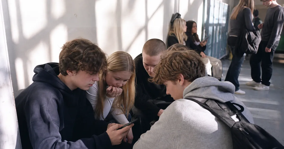 Tonåringar samlade runt en telefon i en skolkorridor, nyfikna på något de tittar på