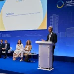 Man i kostym står vid talarstol på konferensbil. På scenen bakom honom sitter sex personer. Blå bakgrund med logotyp för Ukraine Recovery Conference London 2023