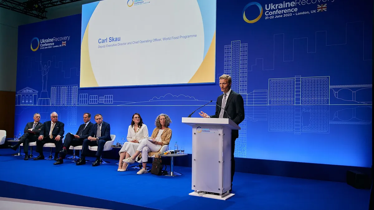 Man i kostym står vid talarstol på konferensbil. På scenen bakom honom sitter sex personer. Blå bakgrund med logotyp för Ukraine Recovery Conference London 2023