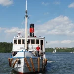 Vitt passagerarfartyg på Mälaren med Drottningholms slott skymtande vid horisonten