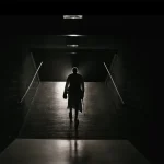 Person i mörk kläder springer genom en lång, mörkläggd tunnel med spotlights uppifrån. Minimalistisk atmosfär med dramaturgi.